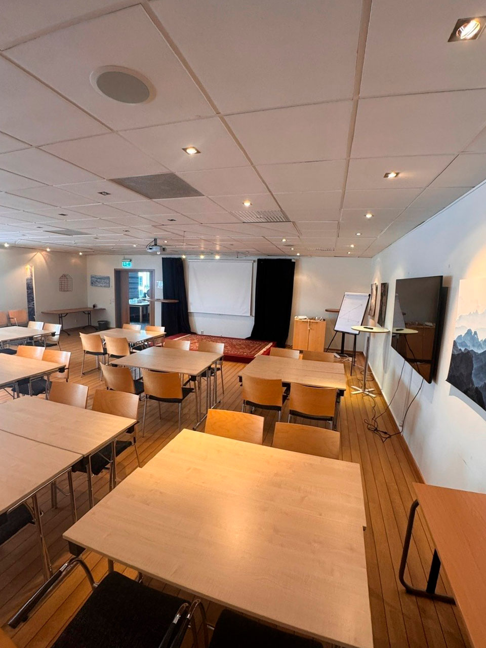 Konferensrum med vackert trägolv och vita väggar, fyllt med bord och stolar.